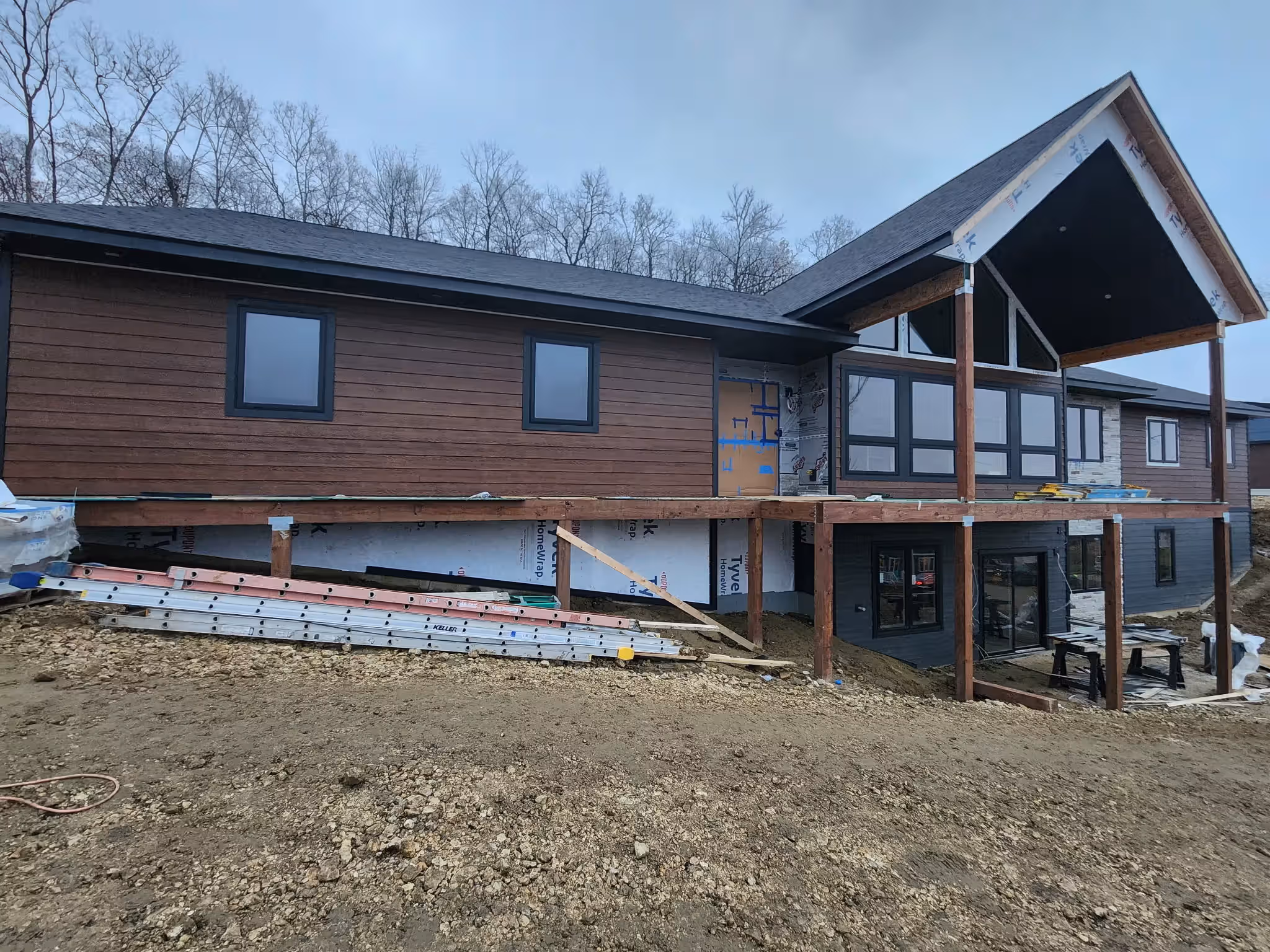 Modern house under construction with wood siding, large windows, and an elevated porch framework.