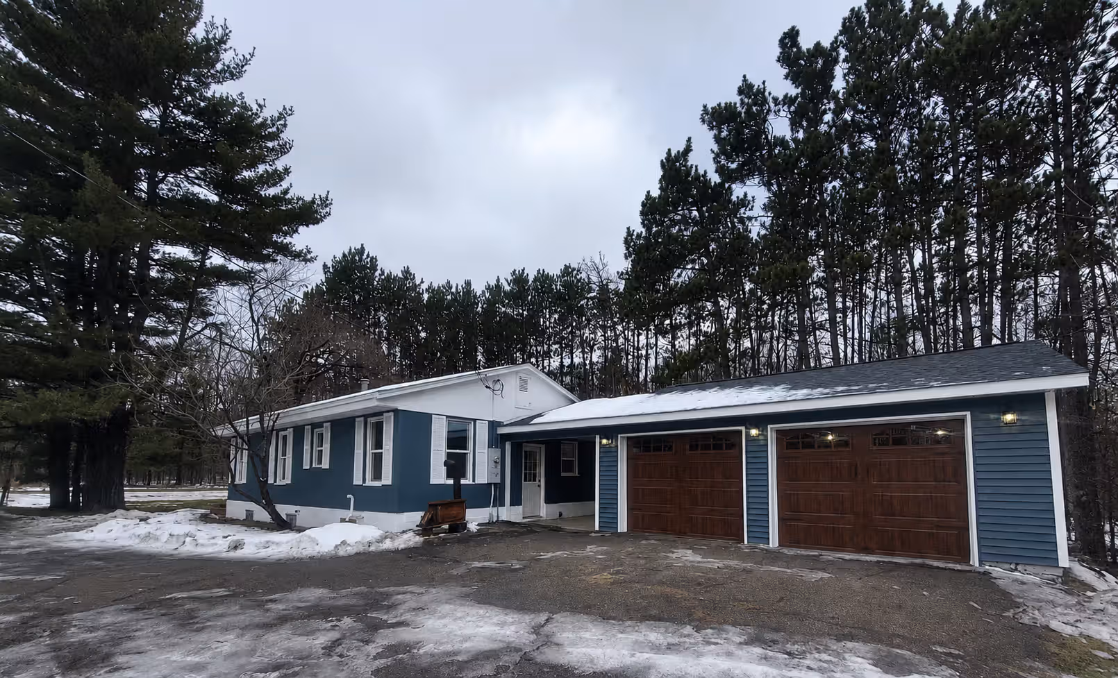 Blue house with white trim and two-car garage with wooden doors on a snowy driveway.